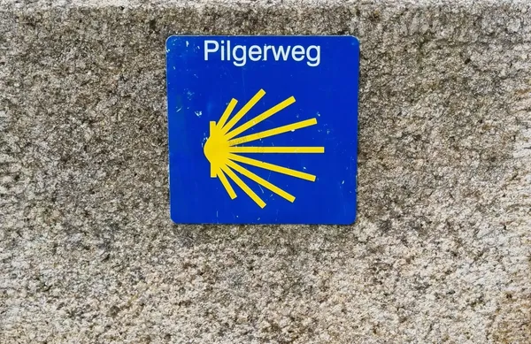 Man sieht einen grauen Steinblock und darauf das blau-gelbe Jakobsweg Zeichen mit der Aufschrift Pilgerweg